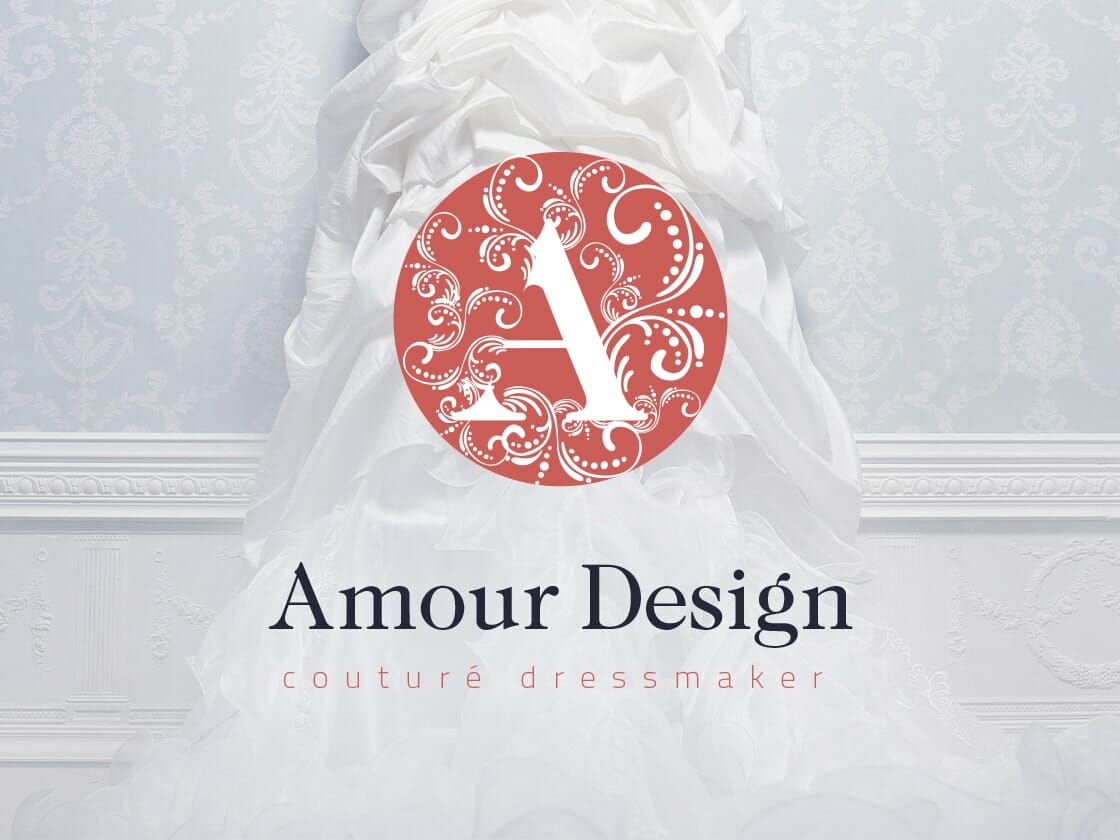 A flowing white couture dress cascades down a light blue patterned wall with intricate white frame detailing. Overlaid in the center is a decorative red emblem featuring an ornate letter "A". Below this emblem, the text reads "Amour Design" with "couturé dressmaker" in smaller type beneath.