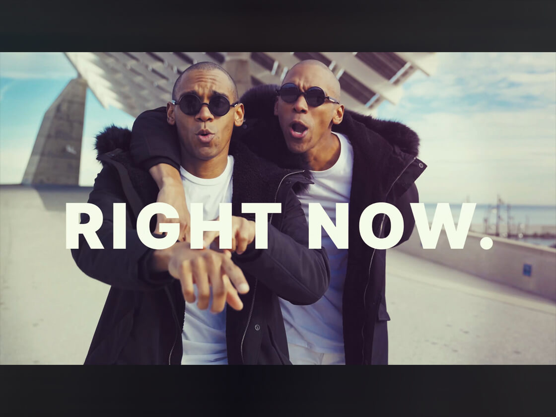 Two men wearing sunglasses and black jackets stand side by side outdoors with one arm around the other's shoulders. Both point forward, and large white text across the image reads "RIGHT NOW." A modern structure and blue sky are in the background.