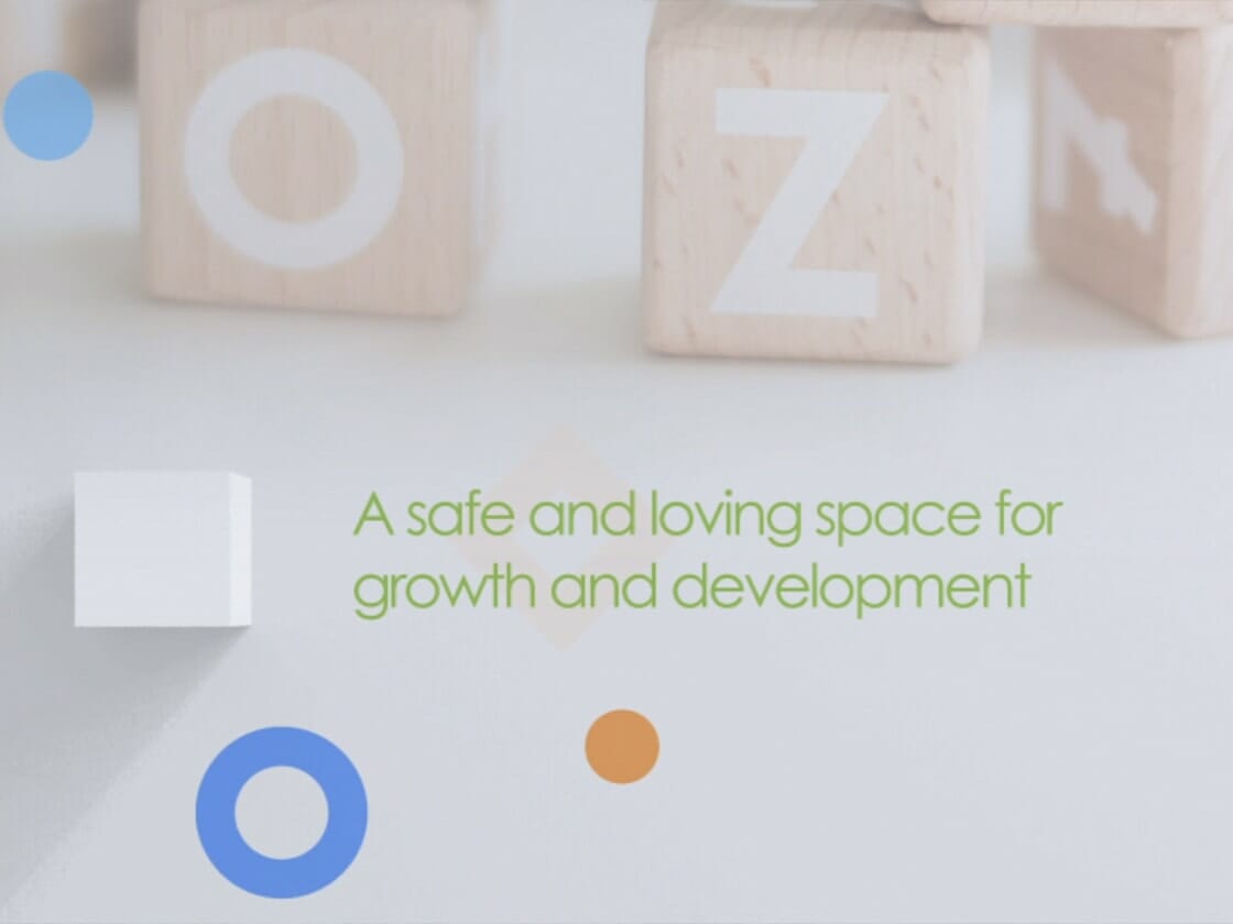Wooden alphabet blocks featuring various letters and shapes are scattered on a white surface. The phrase "A safe and loving space for growth and development" is centered in green text. Colorful circles and geometric shapes in blue, orange, and white frame the text in this charming Oak Grove video opener.