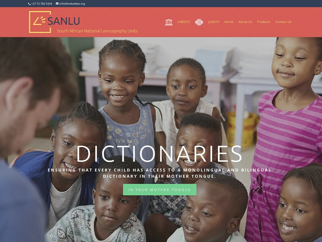 A smiling group of young children gather around a bearded man in a blue shirt as he engages with them. The header on the SANLU website features the logo and name "SANLU" with contact information. The banner reads "DICTIONARIES: Ensuring that every child has access to a monolingual and bilingual dictionary in their mother tongue.