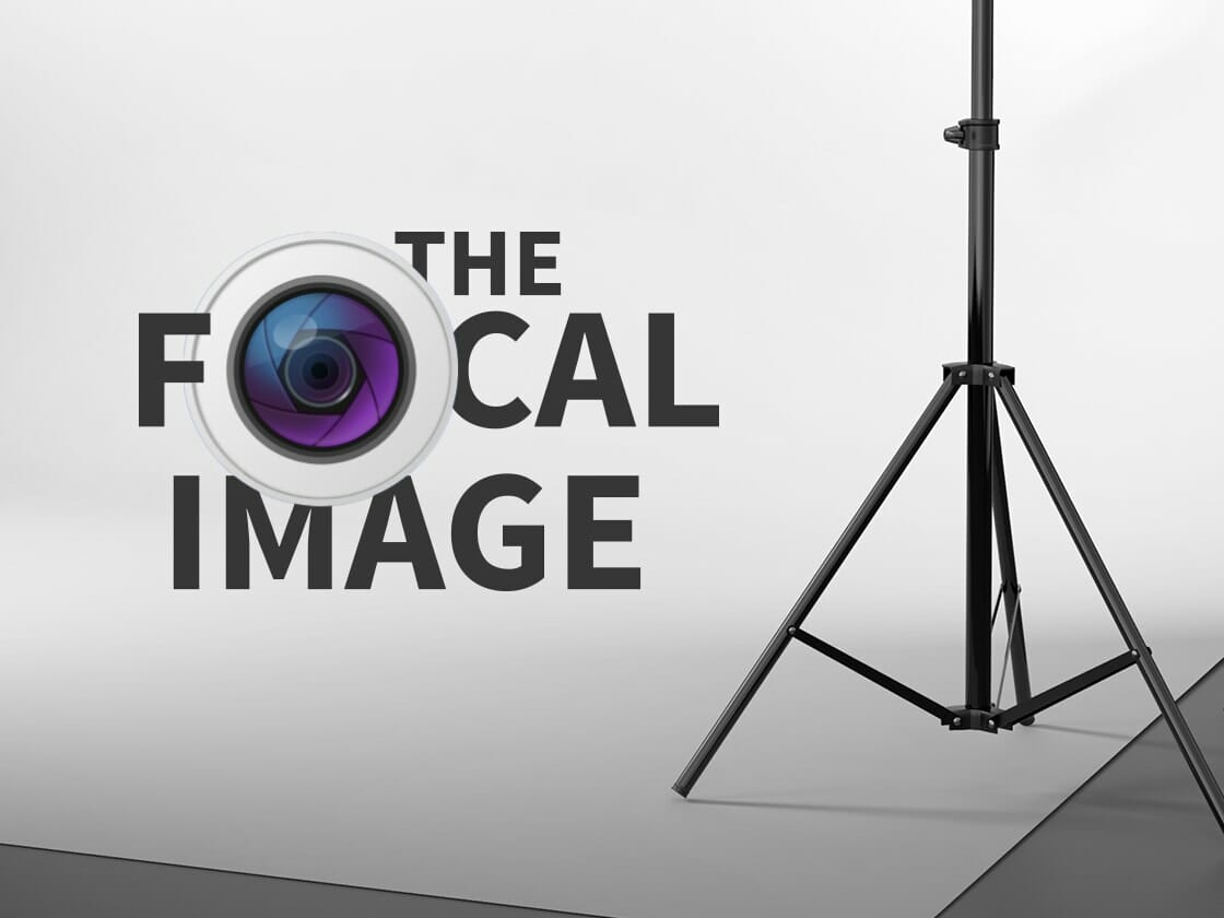 A tripod stand is partially visible on the right side of a white and grey studio backdrop. On the left, bold black capital letters spell out "THE FOCAL IMAGE," with an image of a camera lens replacing the "O" in "FOCAL." The lens features purple and blue gradients.