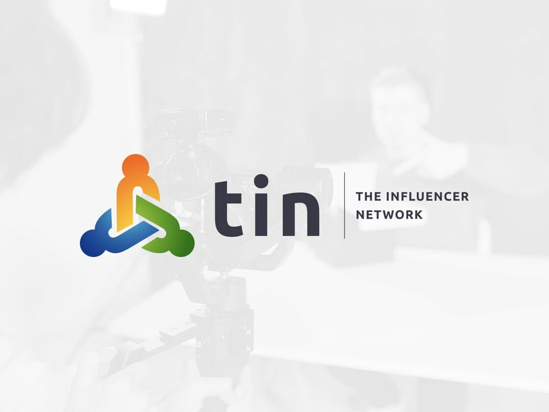 The image features the logo of tin, The Influencer Network, displayed prominently on a white background. The logo includes a multicolored abstract figure and the text "tin" beside it, followed by "The Influencer Network". In the background, a blurred figure and camera equipment are visible.