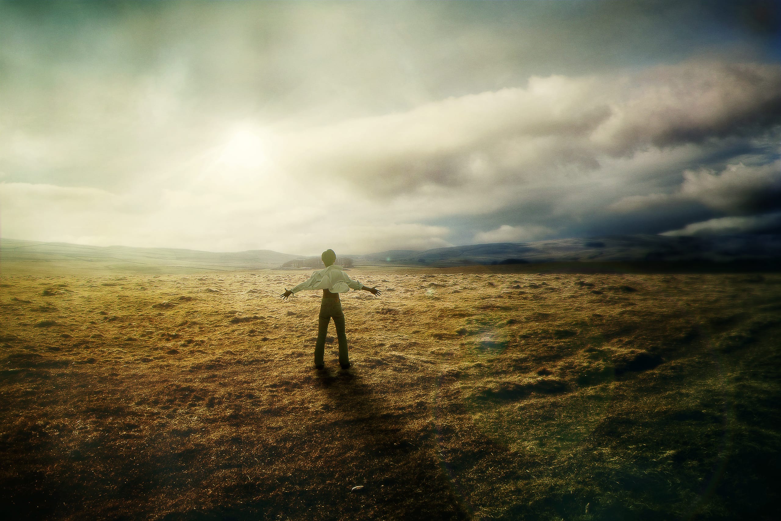 A person standing in an open field with arms outstretched, facing the horizon. The landscape is bathed in warm, golden light, with a dramatic sky filled with clouds. The sun is partially visible, casting a serene glow over the scene, evoking a sense of freedom and tranquility.