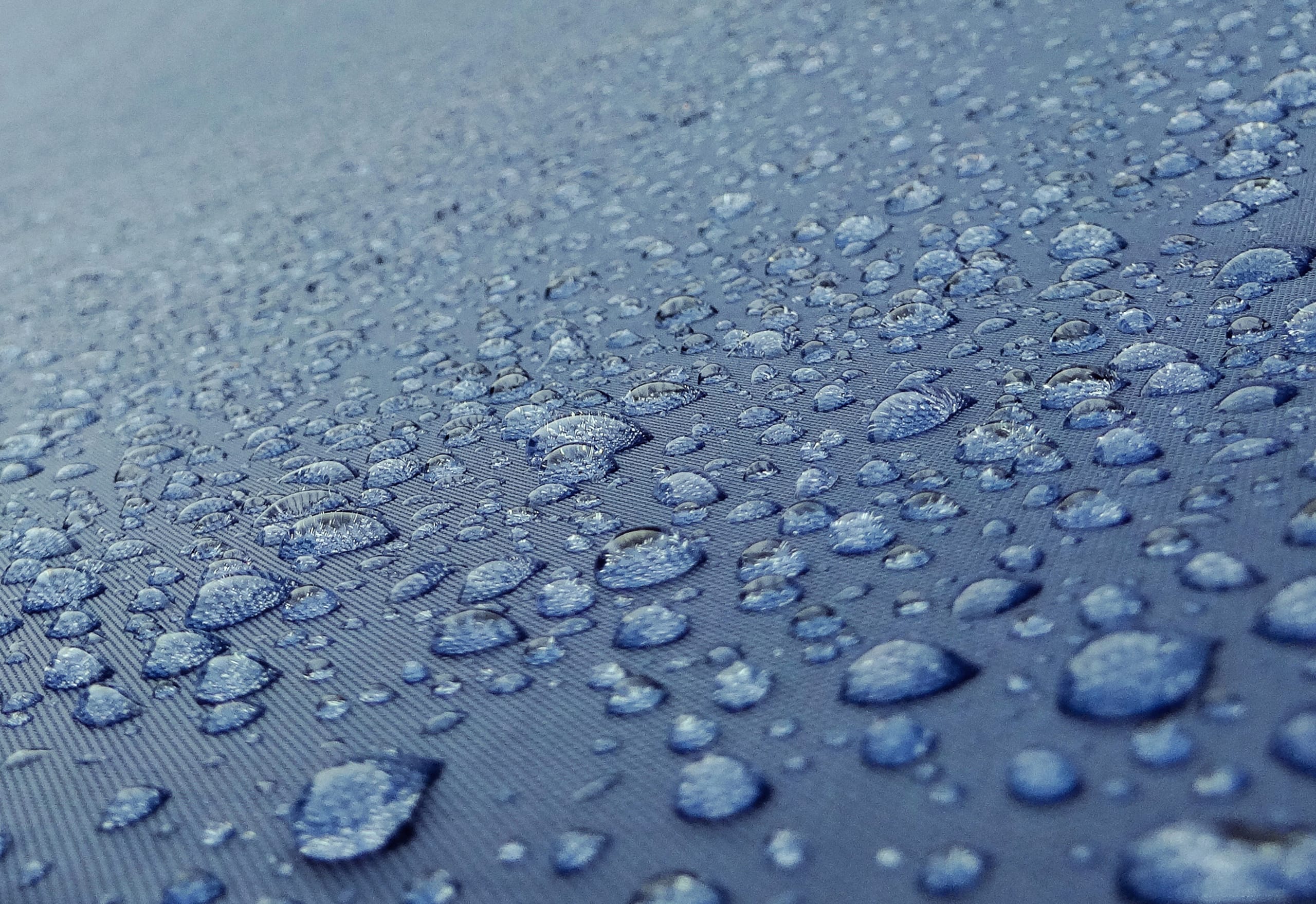 Close-up of numerous water droplets on a blue, textured surface, arranged unevenly and varying in size. The surface appears to be slightly curved, suggesting a three-dimensional form. The droplets glisten, reflecting light, creating a fresh and glossy appearance.