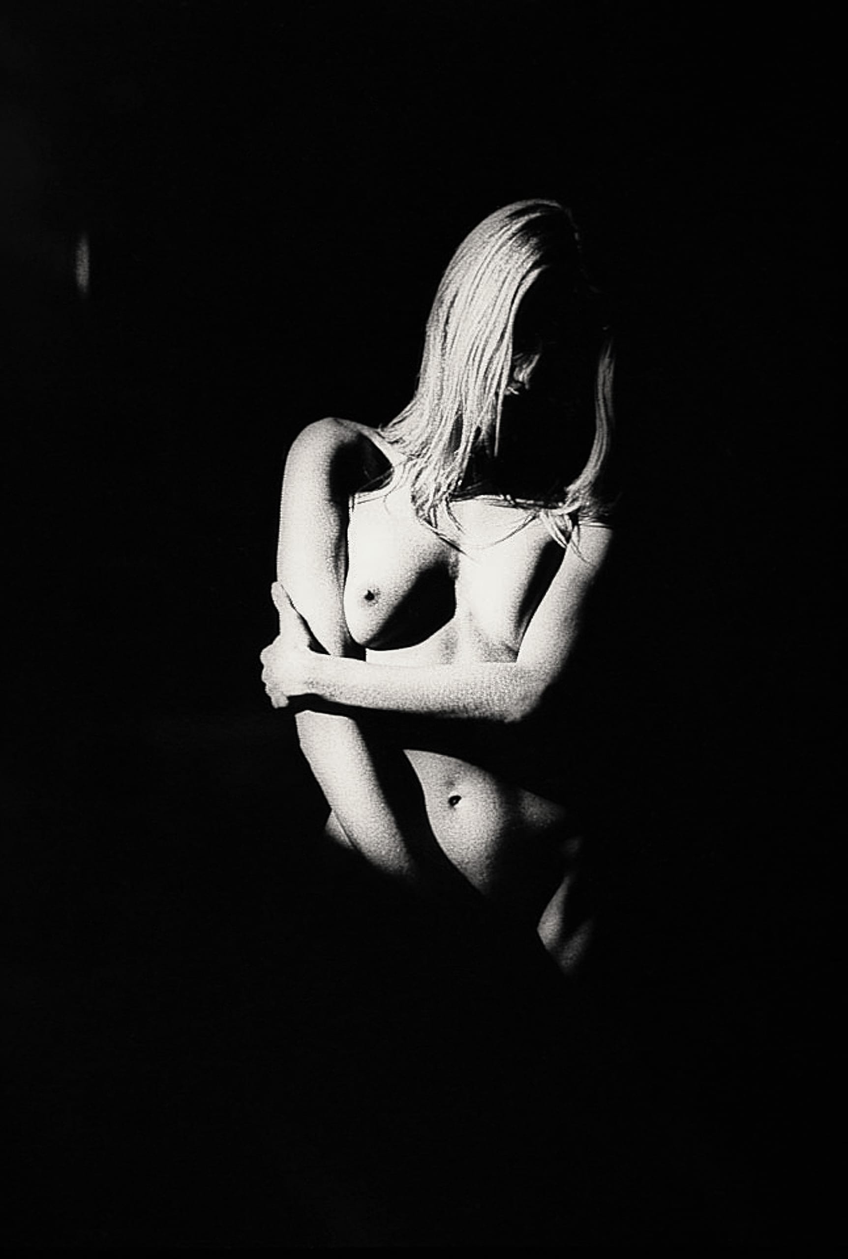 A black and white photograph of a nude person with long hair. They are sitting with their arms crossed over their knees, partially obscured. The image is dramatically lit, casting deep shadows and highlighting the contours of their body, creating a moody, artistic effect.