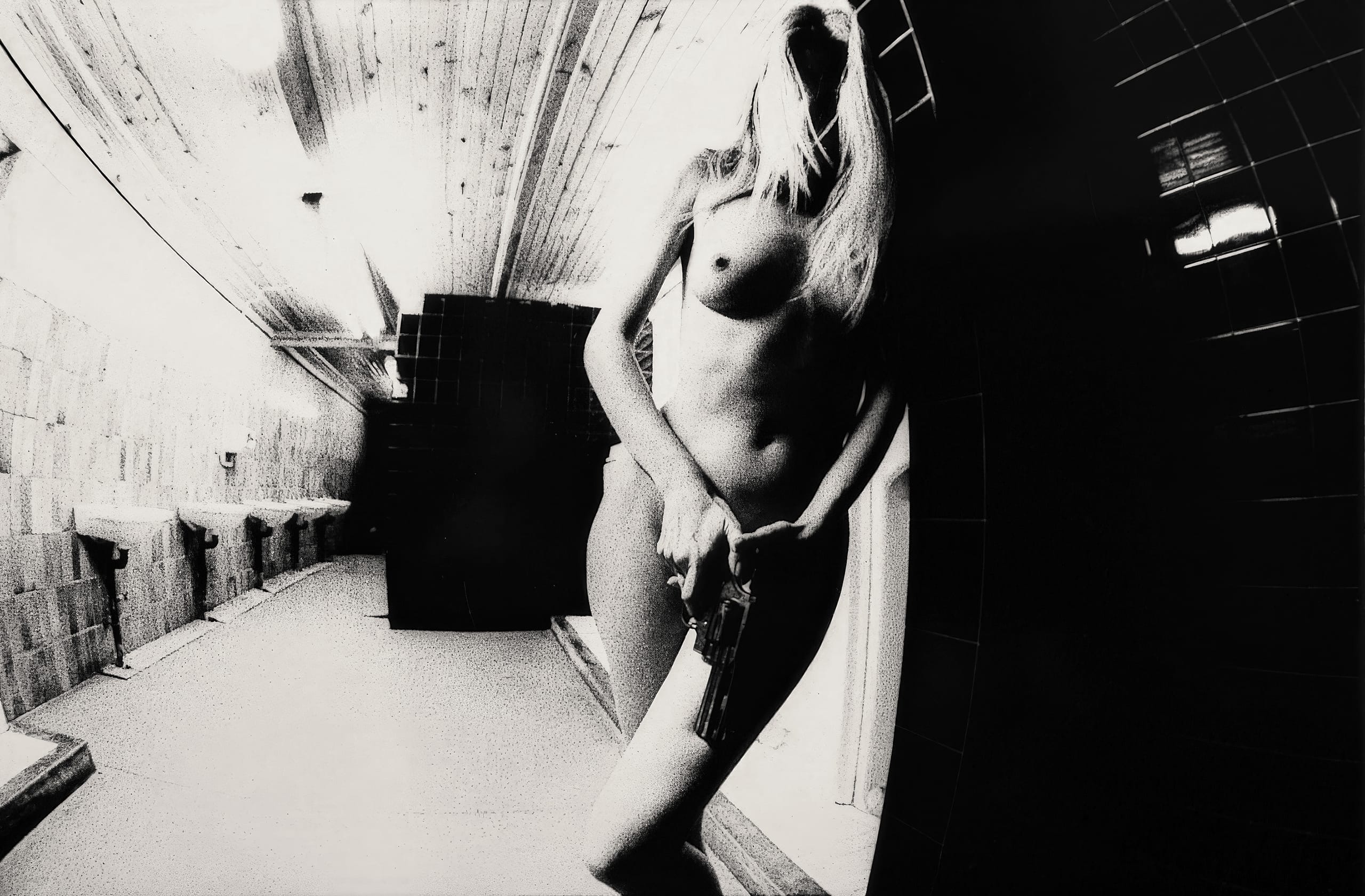Black and white image of a nude person standing in a narrow, dimly lit hallway with tiled walls and a wooden ceiling. The person, partially obscured by shadows, holds a gun by their hip. The perspective is tilted, adding a dramatic and intense atmosphere to the scene.