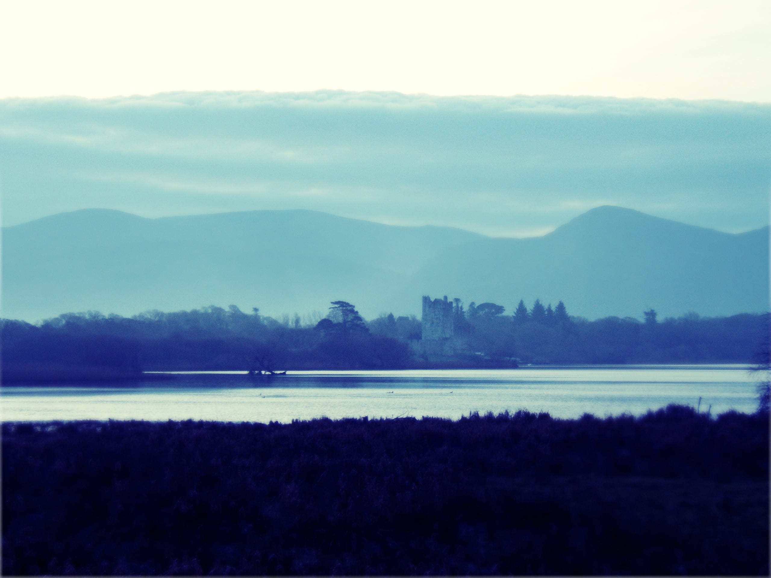A misty blue-toned landscape features a serene lake in the foreground with tall, dark grasses at the water's edge. In the distance, a historic stone castle, evocative of Ireland's rich heritage, sits on a small hill surrounded by dense trees. Behind the castle, layers of rolling hills and mountains are partially obscured by clouds.