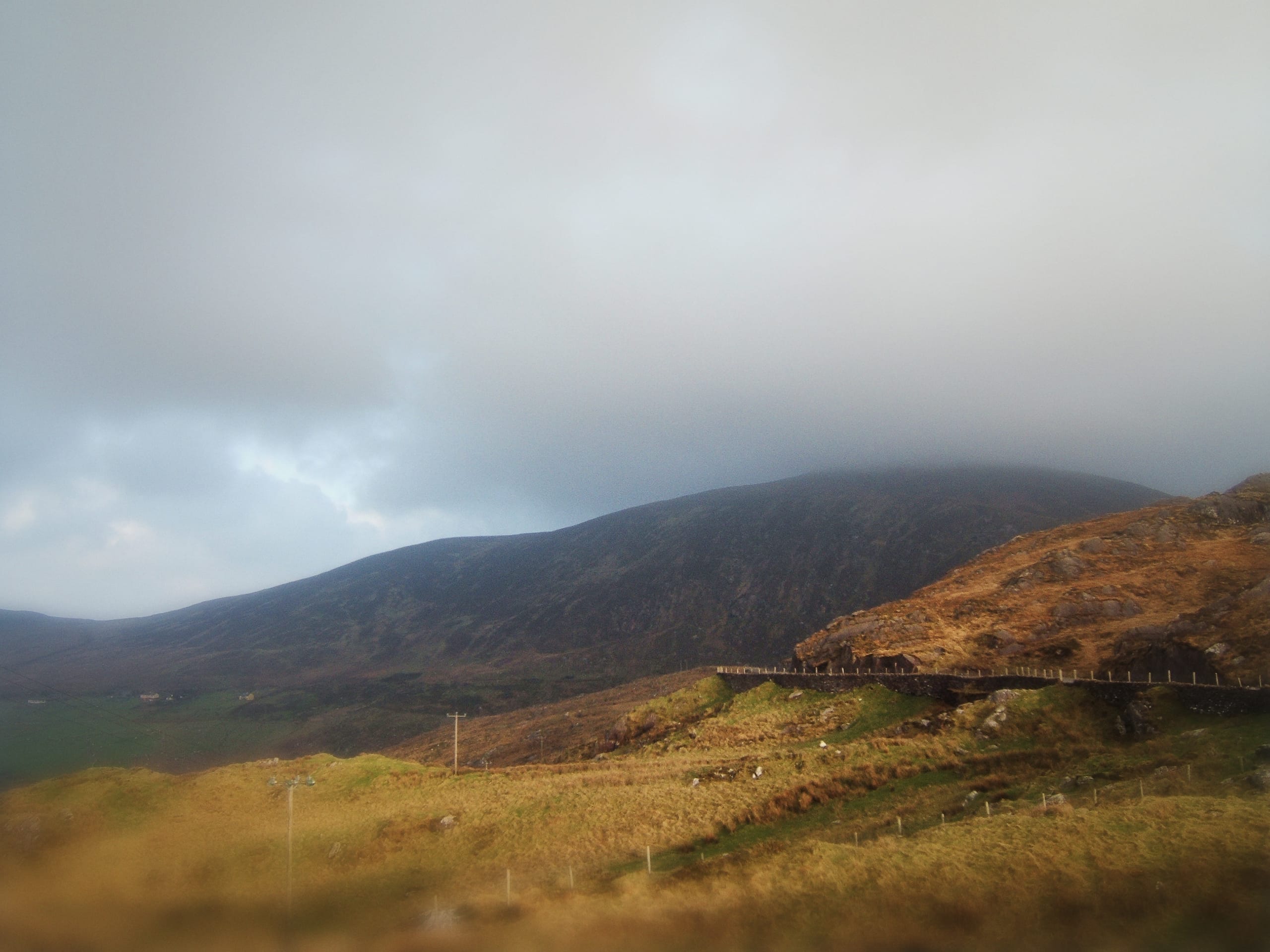 A misty mountain landscape under a cloudy sky. The scene features grassy hills in the foreground, a winding fence along a trail. The slopes of a large, dark mountain veiled in fog dominate the background. This serene route through Ireland captures muted colors and soft lighting.