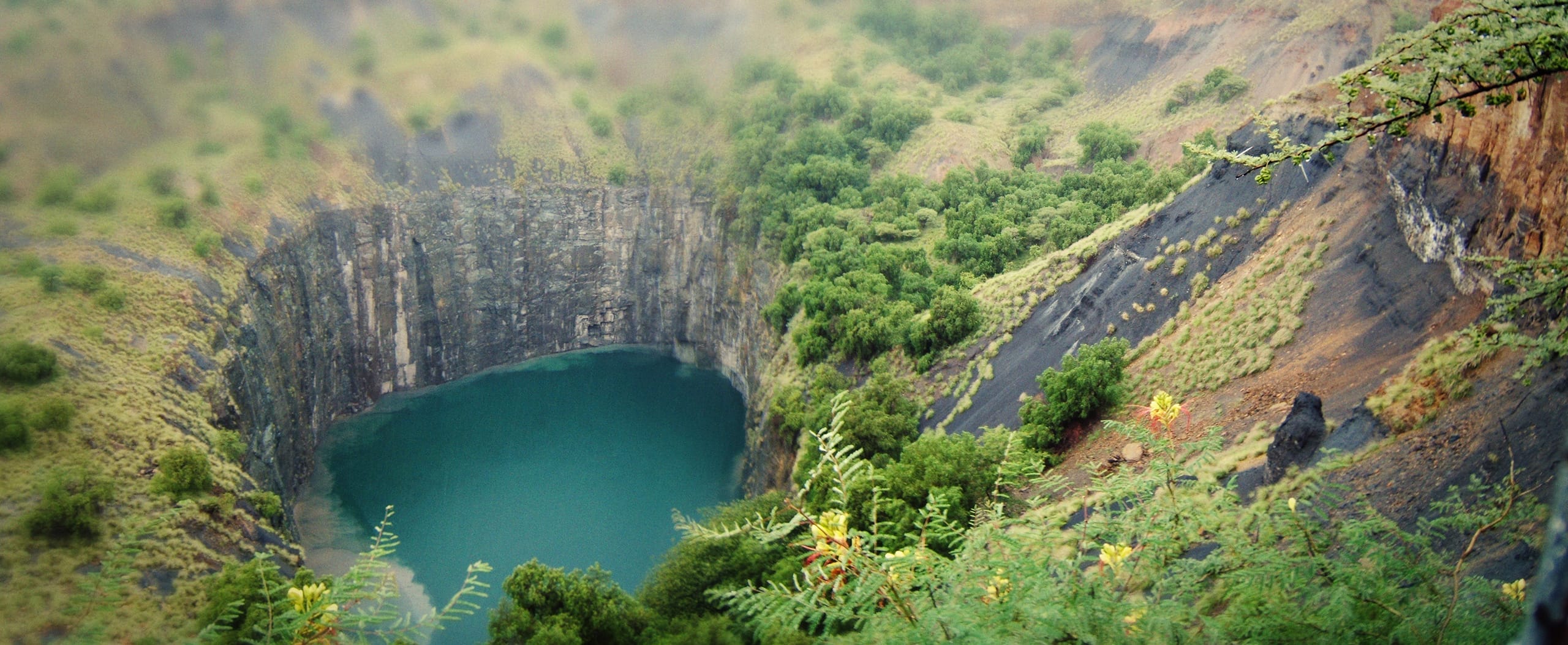 An aerial view of the Big Hole in Kimberley, South Africa, reveals a deep, circular pit with steep rocky walls filled with serene blue-green water. The surrounding area is lush with greenery, scattered trees, and bushes growing on the rocky landscape under an overcast sky.