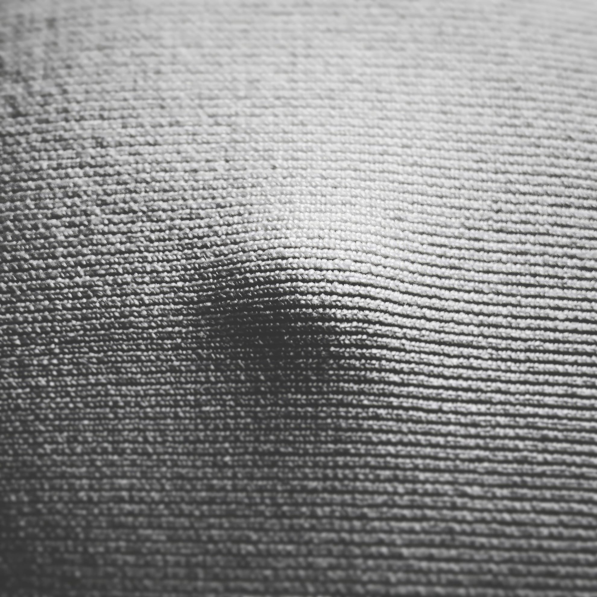 Close-up of a textured gray surface with a slight dent in the center. The material appears woven or knit, with horizontal and vertical lines creating a grid-like pattern. Shadows emphasize the indentation, giving the image a tactile, three-dimensional quality.