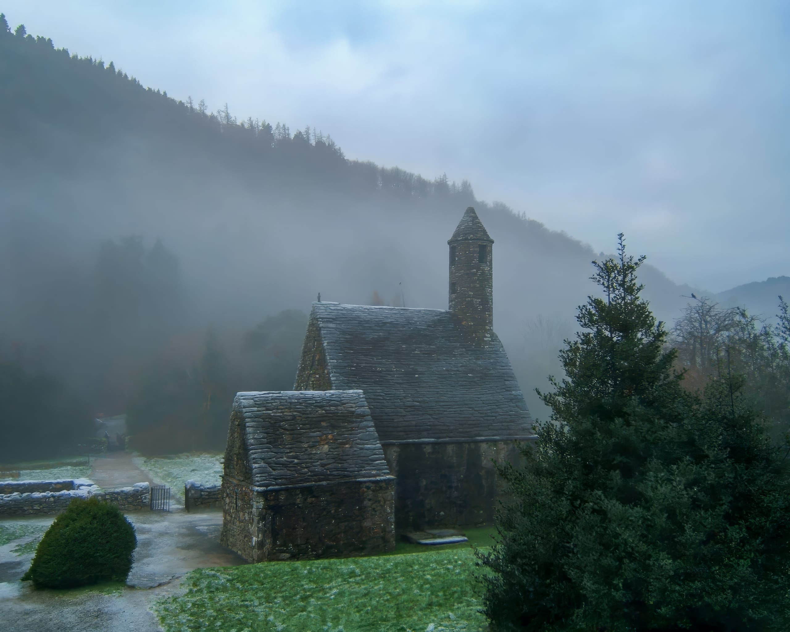 A small, ancient stone church with a tall spire sits surrounded by misty, forested hills. The ground is lightly dusted with frost or snow, and a path leads to the church. Dense evergreen trees and fog add to the serene and mysterious atmosphere of the scene.