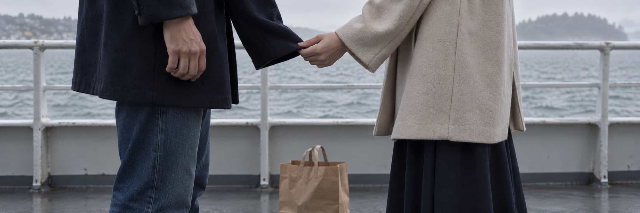 A hand catches the cuff of another person’s sleeve on a ferry, with a paper bag between their shoes.
