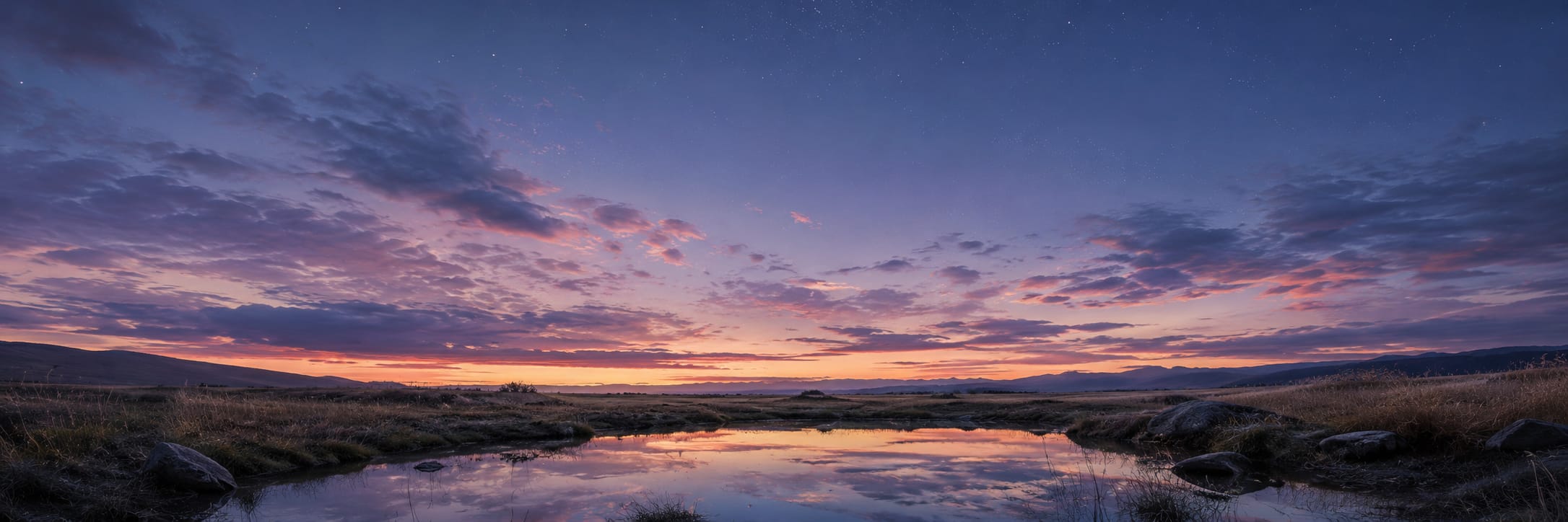 A calm river reflects a sunset sky over open grassy land, distant hills, and scattered clouds.