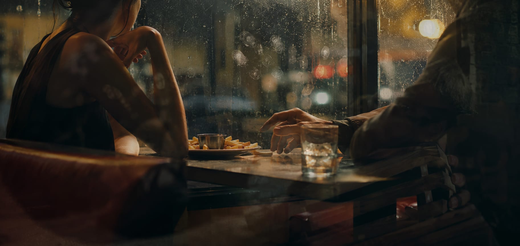 Two people share fries at a table by a rainy window in a quiet restaurant.