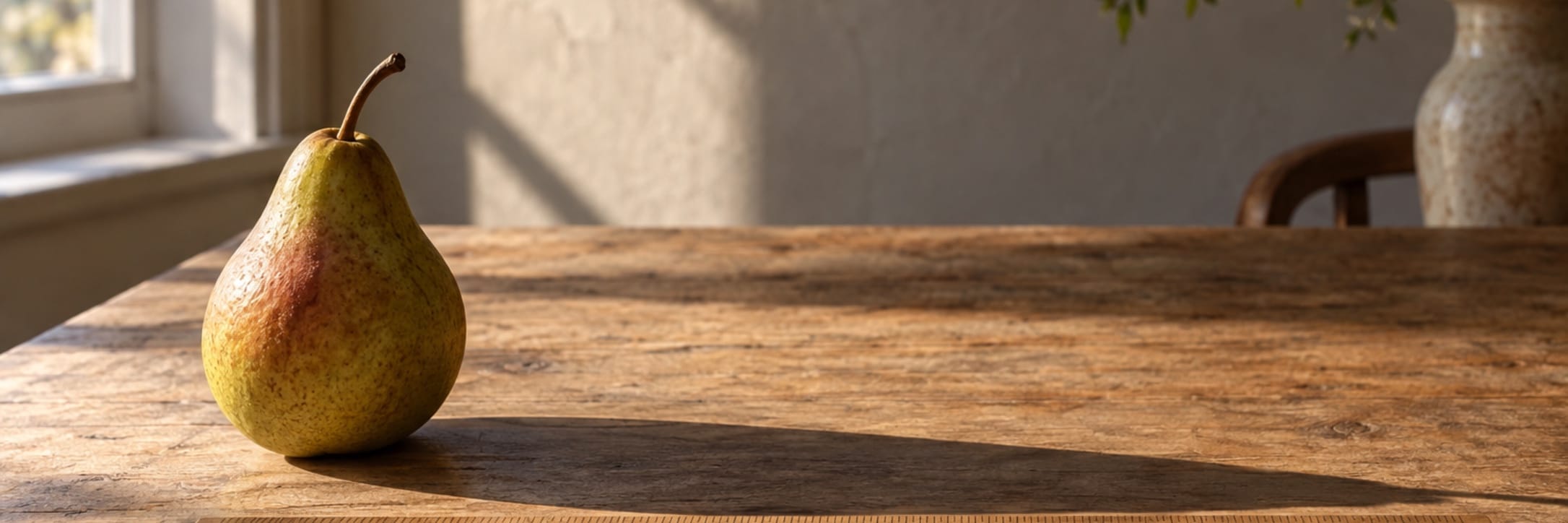 A pear casts a long shadow across a wooden table in warm sunlight.