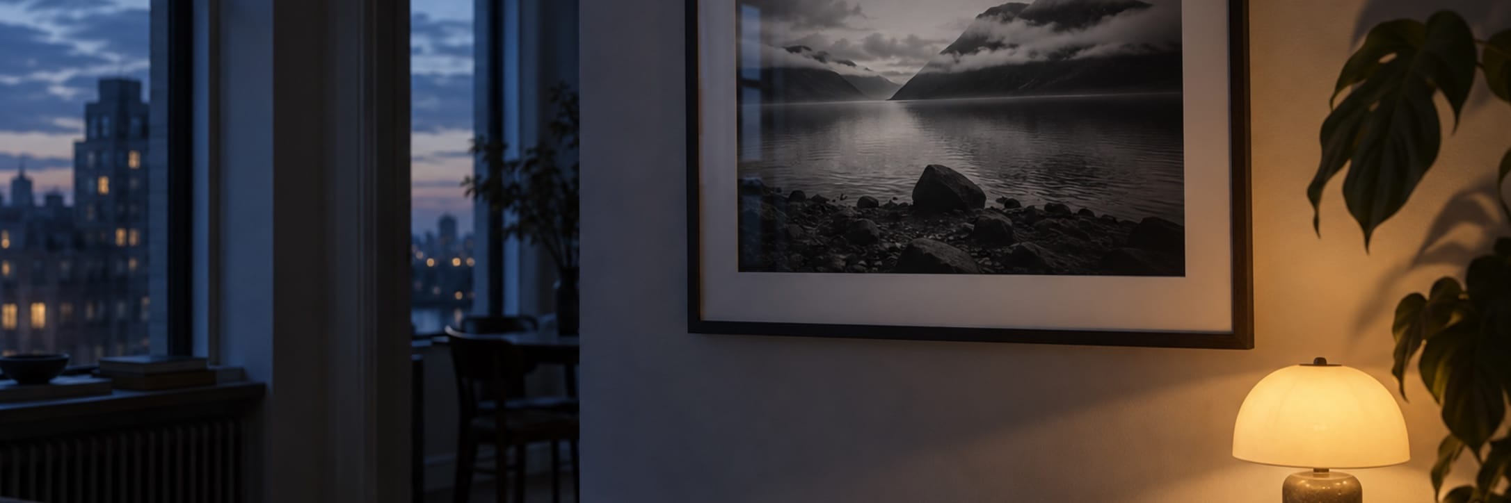 A framed landscape print hangs in a shadowed room beside a glowing lamp and plant.